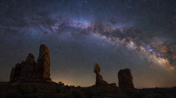 The Milky Way arching over a dark desert landscape with rock formations